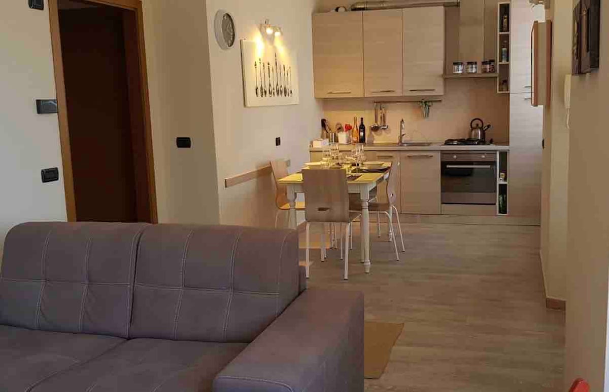 A modern kitchen is visible, featuring light wood cabinetry and stainless steel appliances. A dining table is set for four, accompanied by a cozy sitting area that includes a gray couch. Warm lighting enhances the inviting atmosphere of the open floor plan.