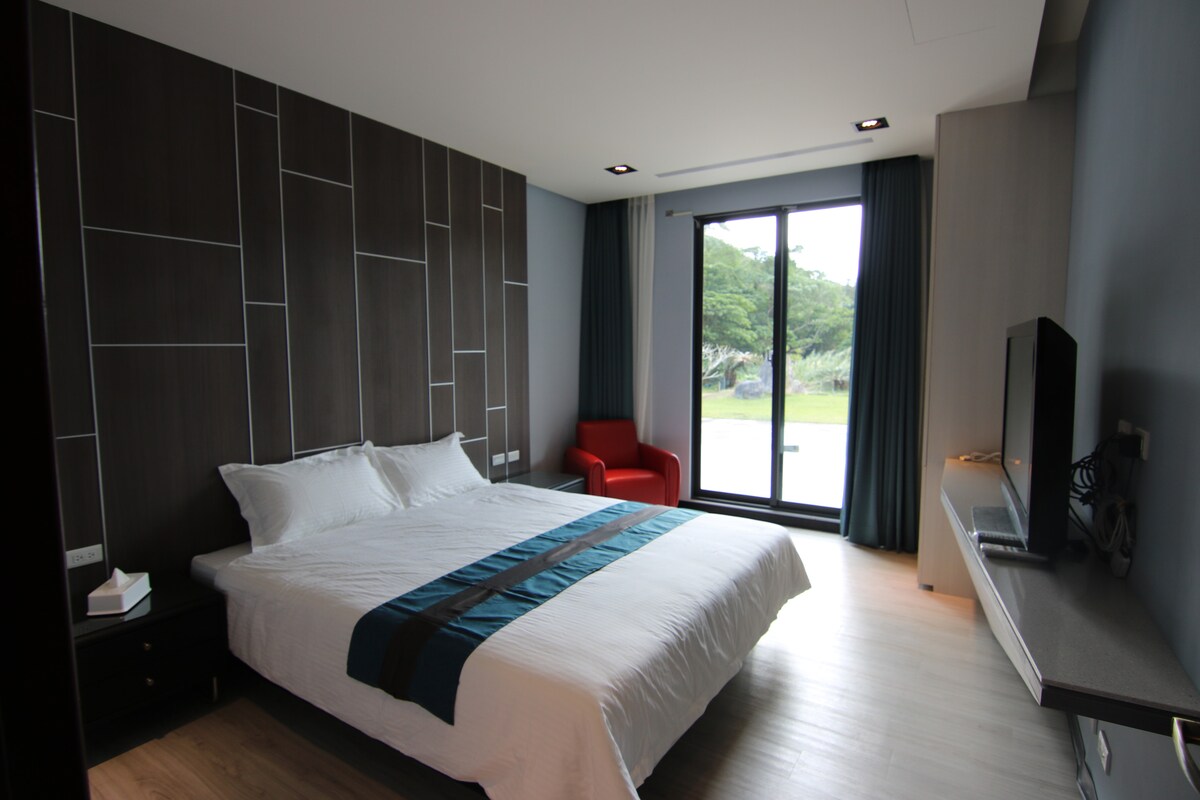 A well-lit bedroom features a spacious bed with a decorative blanket and a modern accent wall. A comfortable red chair is positioned beside a large window, offering a view of the green outdoor space. A television is mounted on the wall opposite the bed.