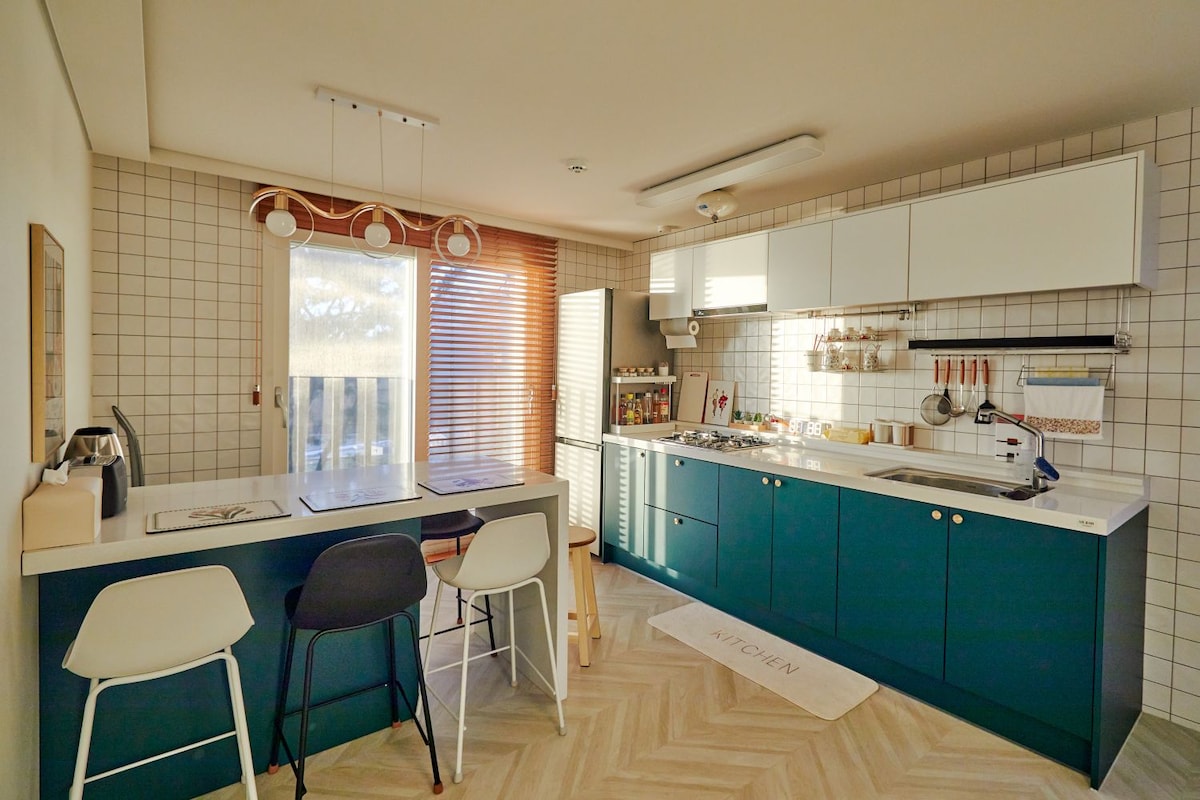 The modern kitchen features teal cabinets and a light-colored countertop. A dining area with two stools is positioned near the window. Natural light streams through the window, accentuating the tiles and utensils organized neatly along the walls.