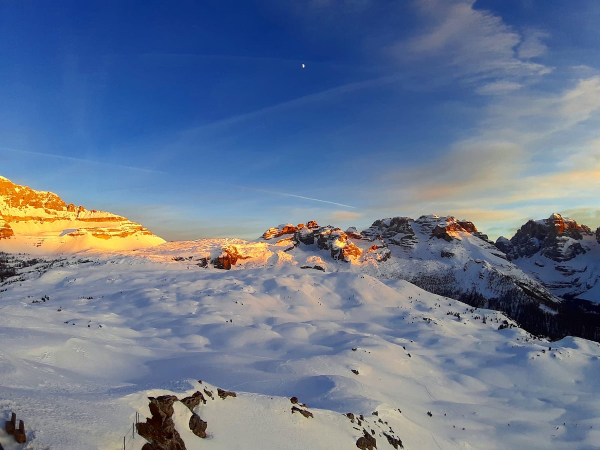 Snow-covered mountains are illuminated by the soft glow of the setting sun, creating a contrasting palette of warm and cold hues. The landscape's undulating terrain is visible, with a clear sky adding to the serene atmosphere of the Dolomites.