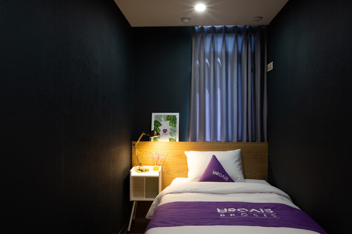 A cozy bedroom features dark walls and is illuminated by soft lighting. A spacious bed with white linens and a purple accent pillow is positioned against a wooden headboard. A small bedside table holds a lamp, and a window with sheer curtains allows natural light to filter in.