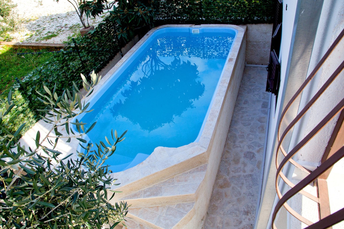 An outdoor private pool, surrounded by greenery, captures clear blue water. The pool features a textured stone edge and steps gracefully leading into the water. Natural light enhances the serene atmosphere of the space with a view of lush foliage nearby.
