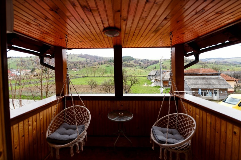 A cozy terrace features two hanging chairs with comfortable cushions, positioned around a small table. The space is surrounded by wooden walls and offers panoramic views of the lush green landscape and distant hills.