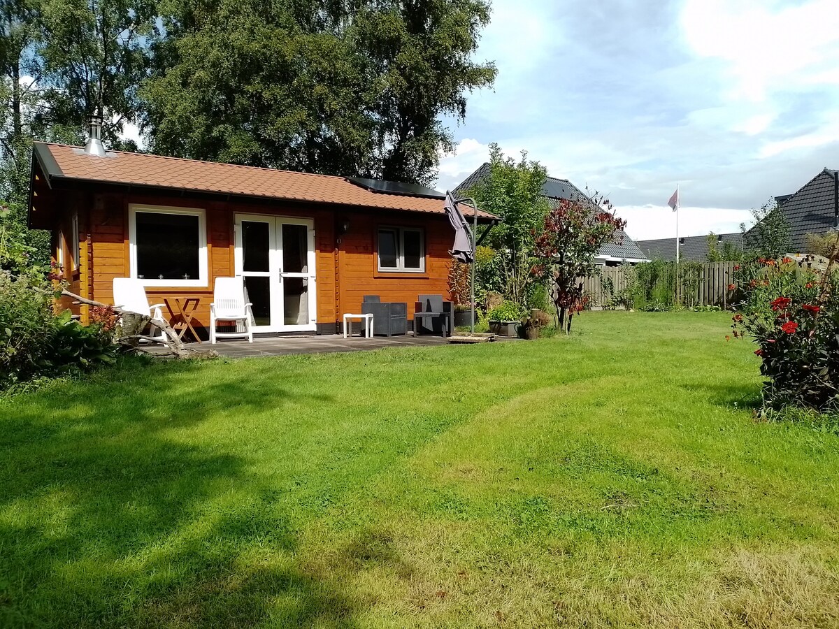 A charming wooden cabin is set against a backdrop of green grass and lush trees. A patio area features two white chairs and a small seating arrangement. The surrounding garden includes flower beds, enhancing the peaceful outdoor environment.
