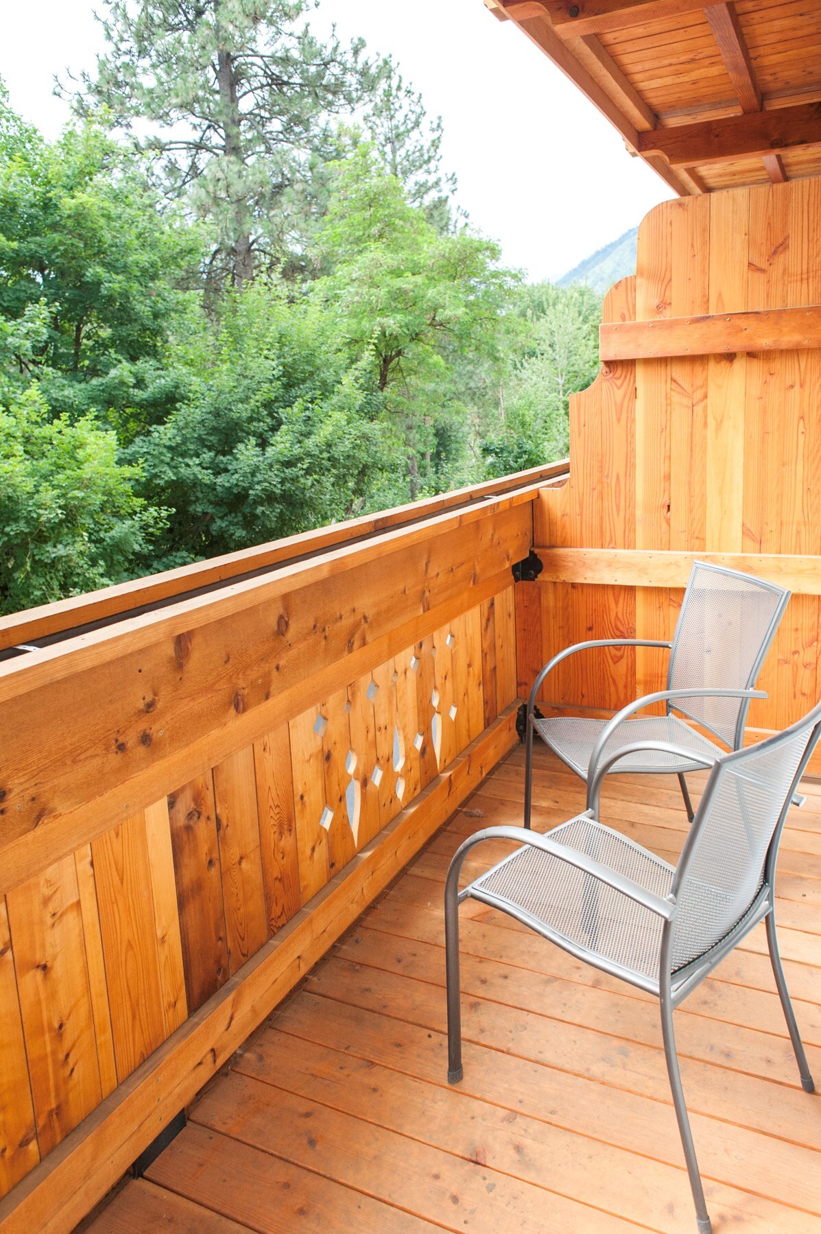 A private balcony features wooden paneling and a natural wood floor. Two metal chairs are positioned along the railing, offering views of surrounding trees and greenery. The serene environment promises a peaceful outdoor space for relaxation.