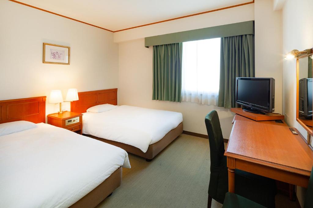 A hotel room featuring two spacious beds with white bedding and a wooden headboard. A window is covered with light green curtains, allowing natural light to enter. A small desk with a chair and a flat-screen television are positioned against one wall.