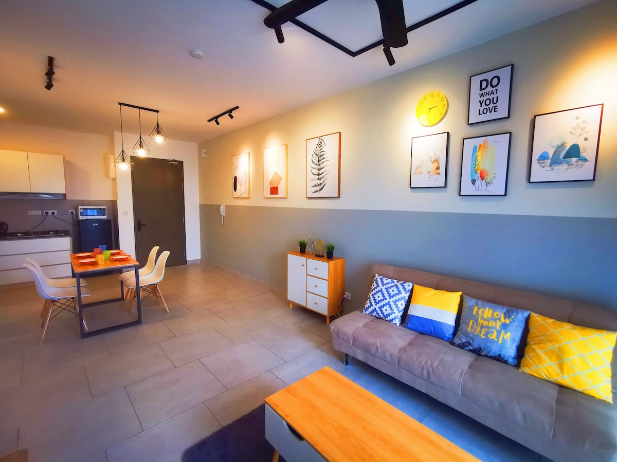 A modern living space features a comfortable sofa adorned with decorative cushions, paired with a wooden coffee table. A dining area with a table and four chairs is visible, while framed artwork adorns the walls, contributing to a bright and inviting atmosphere.
