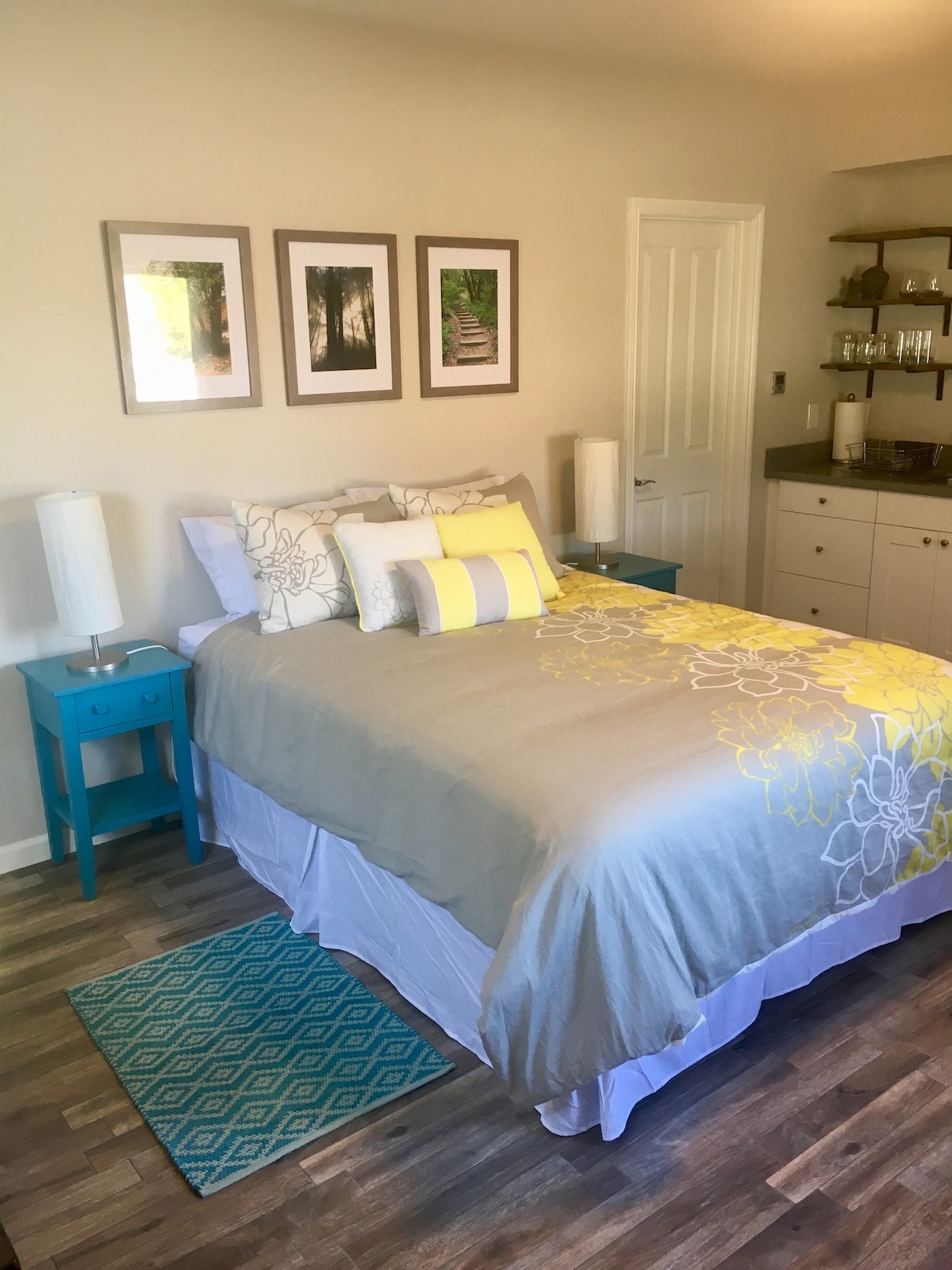 The sleeping area features a spacious bed dressed in gray and yellow linens, complemented by decorative pillows. Light wood floors are showcased beneath a textured area rug. Framed botanical prints hang above the bed, while a compact kitchenette is visible to the side.