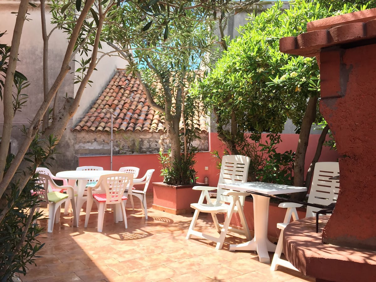 A private garden patio is showcased, featuring several white plastic tables and chairs arranged among greenery. A barbecue area is visible, framed by lush plants and trees. The sunlit space is bordered by red-painted walls and a tiled roof in the background.
