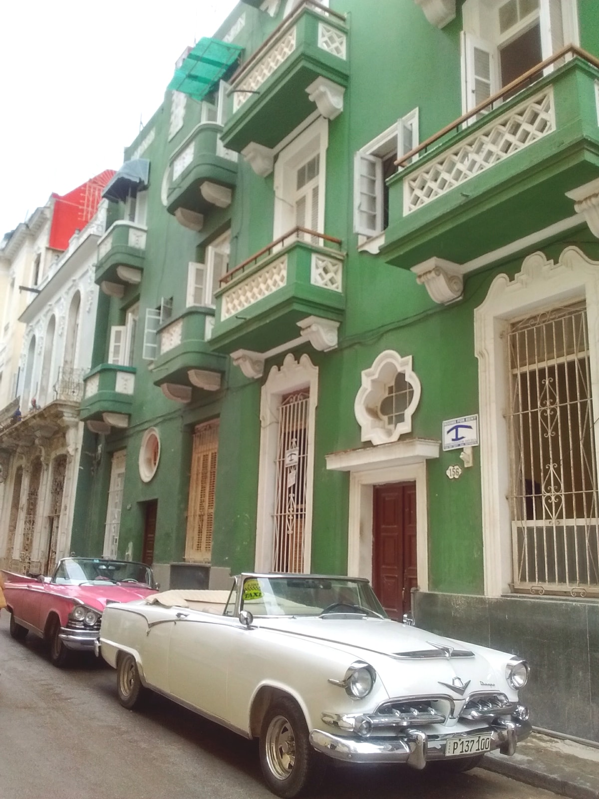 A vibrant green building with ornate balconies stands along a narrow street. Classic vintage cars, including a white convertible and a red one, are parked curbside. The facade displays decorative architectural elements, with laundry hanging from the balconies, adding a touch of local life.