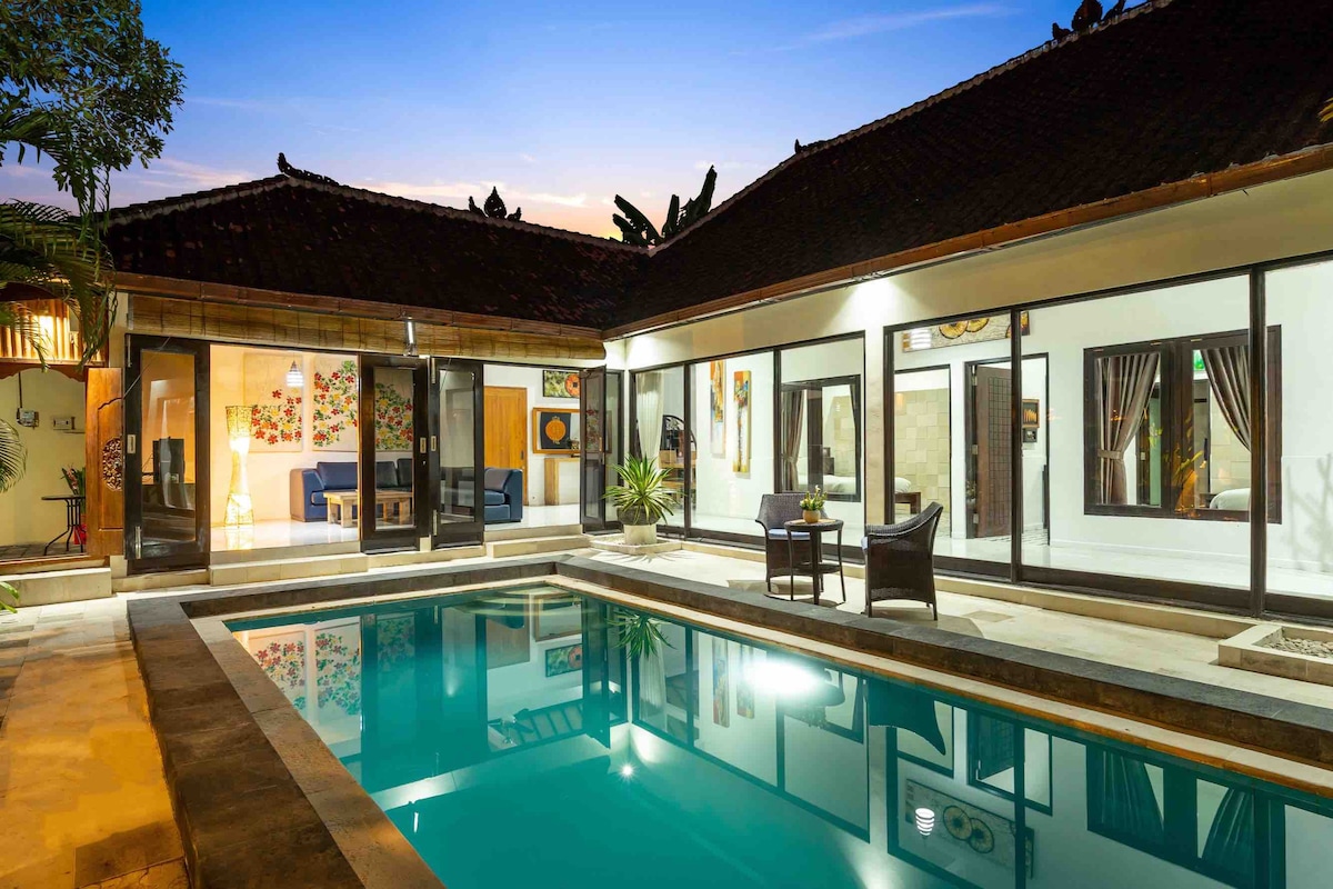 The image captures a tranquil outdoor area featuring a private swimming pool with clear water. Surrounding the pool, large glass doors lead to an indoor lounge area. The space is complemented by tropical plants and evening light illuminating the serene environment.