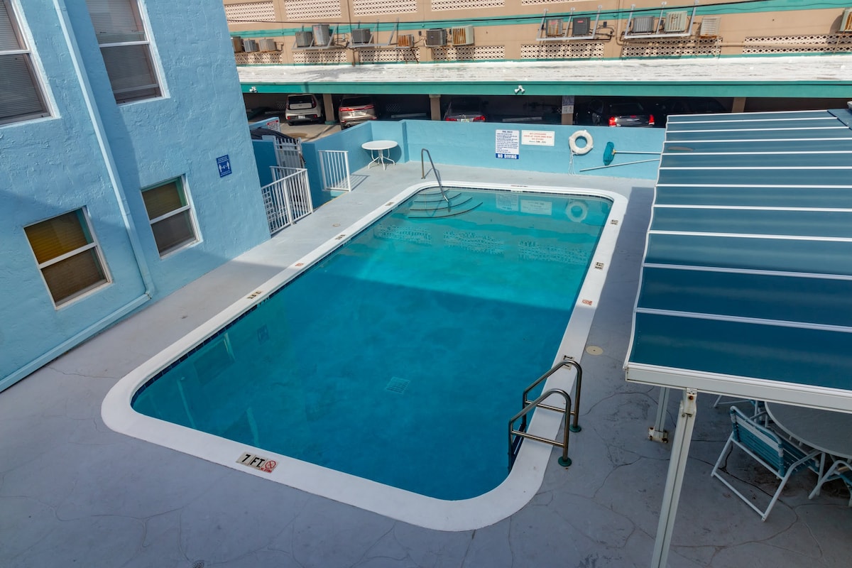 An outdoor pool is depicted, surrounded by a light blue building. The pool's clear water reflects sunlight, and a railing is positioned at one end. A shaded seating area features multiple chairs under a blue awning, with signs of nearby parking visible in the background.
