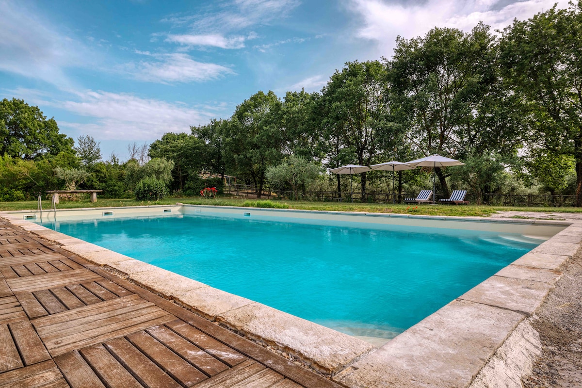A spacious outdoor pool, framed by natural stone and wooden decking, reflects the clear sky. Sun loungers positioned under umbrellas provide a relaxing setting, with greenery observed in the background, enhancing the tranquil environment.