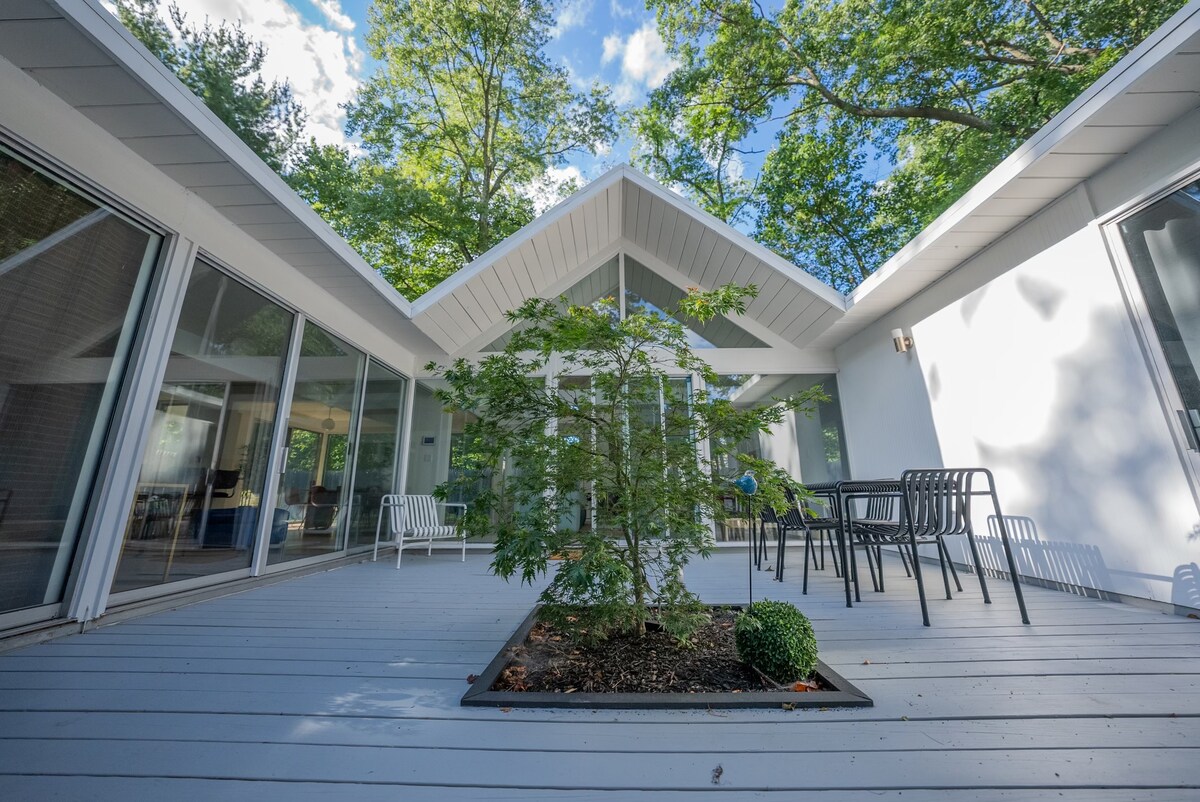 An outdoor space is surrounded by a harmonious arrangement of glass walls, allowing natural light to enrich the area. A Japanese Maple tree is featured in the center, with seating arrangements visible on the perimeter, inviting relaxation under the trees.