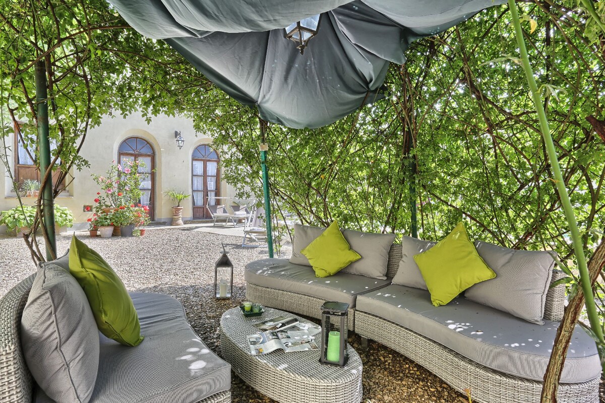 A cozy outdoor seating area is shaded by lush greenery, featuring a curved sectional sofa with soft grey cushions and vibrant green throw pillows. A round table is accompanied by decorative lanterns, creating a restful space for guests to relax amidst the natural surroundings.