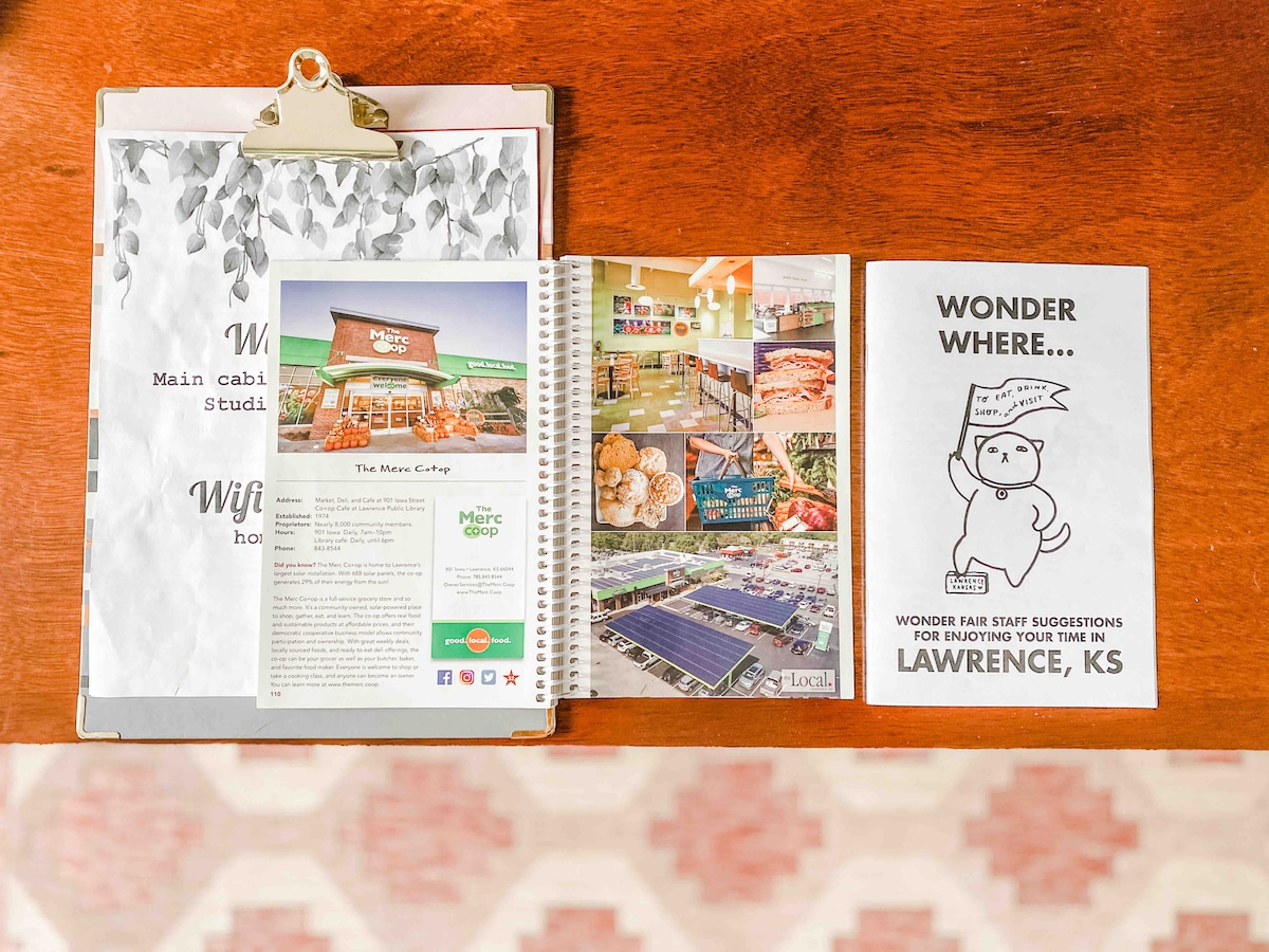 A wooden table is set with a clipboard holding a welcome note. A spiral-bound guidebook displays local dining options, and a pamphlet provides suggestions for activities in Lawrence. The arrangement is complemented by a patterned rug beneath.