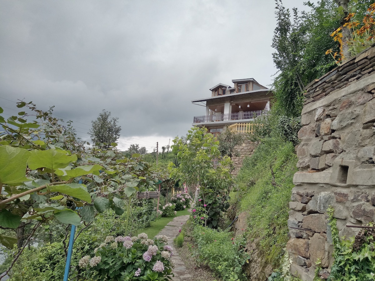 A gently winding stone path leads through lush green gardens, with colorful flowers and foliage lining the way. In the background, a two-story house with a wooden balcony is set against a cloudy sky, surrounded by trees and natural beauty.