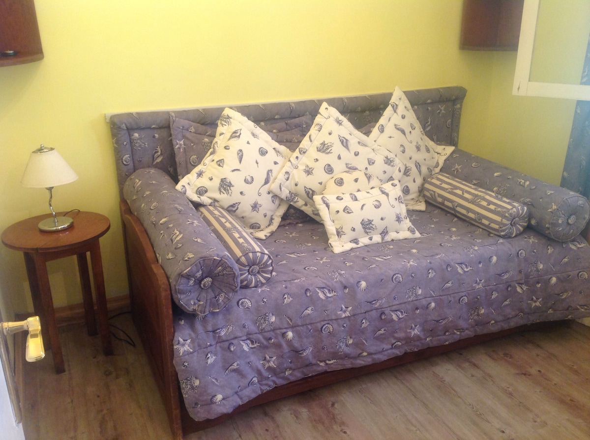 A cozy sleeping area features a daybed adorned with decorative pillows in a marine motif. A small nightstand with a lamp is positioned nearby, and soft yellow walls contribute to the warm ambiance. Natural light is allowed through a nearby window.