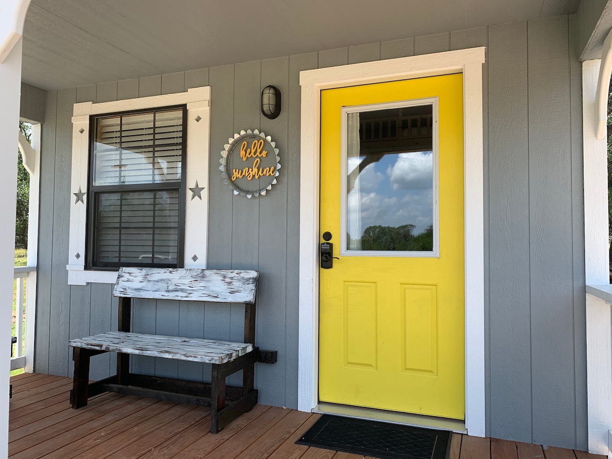 A welcoming porch area is seen featuring a bright yellow door centered within a gray wooden exterior. A rustic bench sits on a wooden deck, accompanied by a decorative circular sign that reads 'Hello Sunshine,' and windows adorned with dark grills allow for natural light.