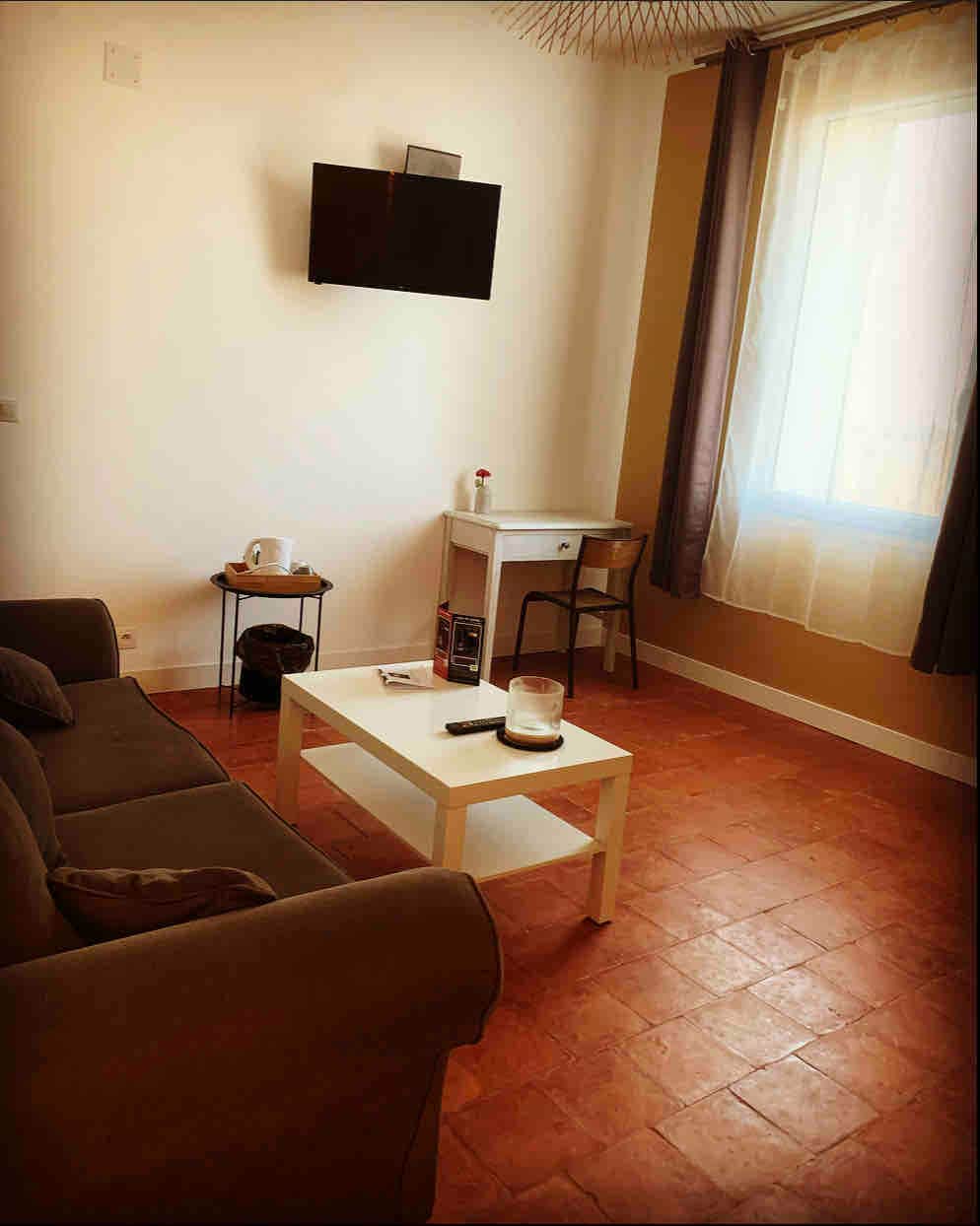 A cozy seating area is furnished with a dark sofa and a small white table. A television is mounted on the wall, while natural light enters through a nearby window, adorned with light curtains. A desk and chair are positioned against the wall.