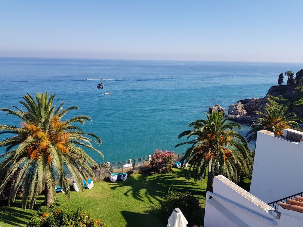 A view of the tranquil Mediterranean Sea, with a boating scene in the distance. Lush palm trees frame the scene, mingling with vibrant flowering plants in the garden. The coastline is visible, alongside sun loungers arranged on the green lawn, creating a peaceful outdoor atmosphere.