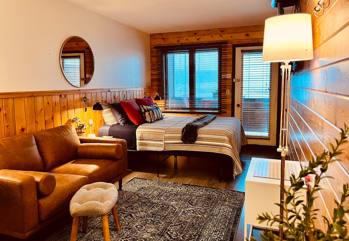 A spacious hotel-style room features warm wooden paneling and large windows offering natural light. A comfortable queen bed is dressed in layered bedding, accompanied by a cozy loveseat and a small stool. A decorative rug enhances the inviting atmosphere, with a small table positioned beside the bed.