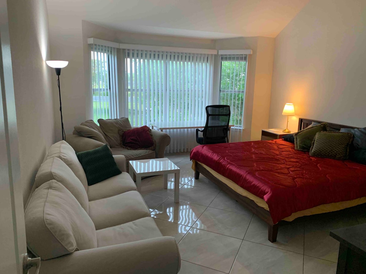 A spacious room features a large bed adorned with a red covering, complemented by decorative pillows. A seating area includes a light-colored couch and a small white coffee table. Natural light filters through vertical blinds of large windows, illuminating the tile floor.