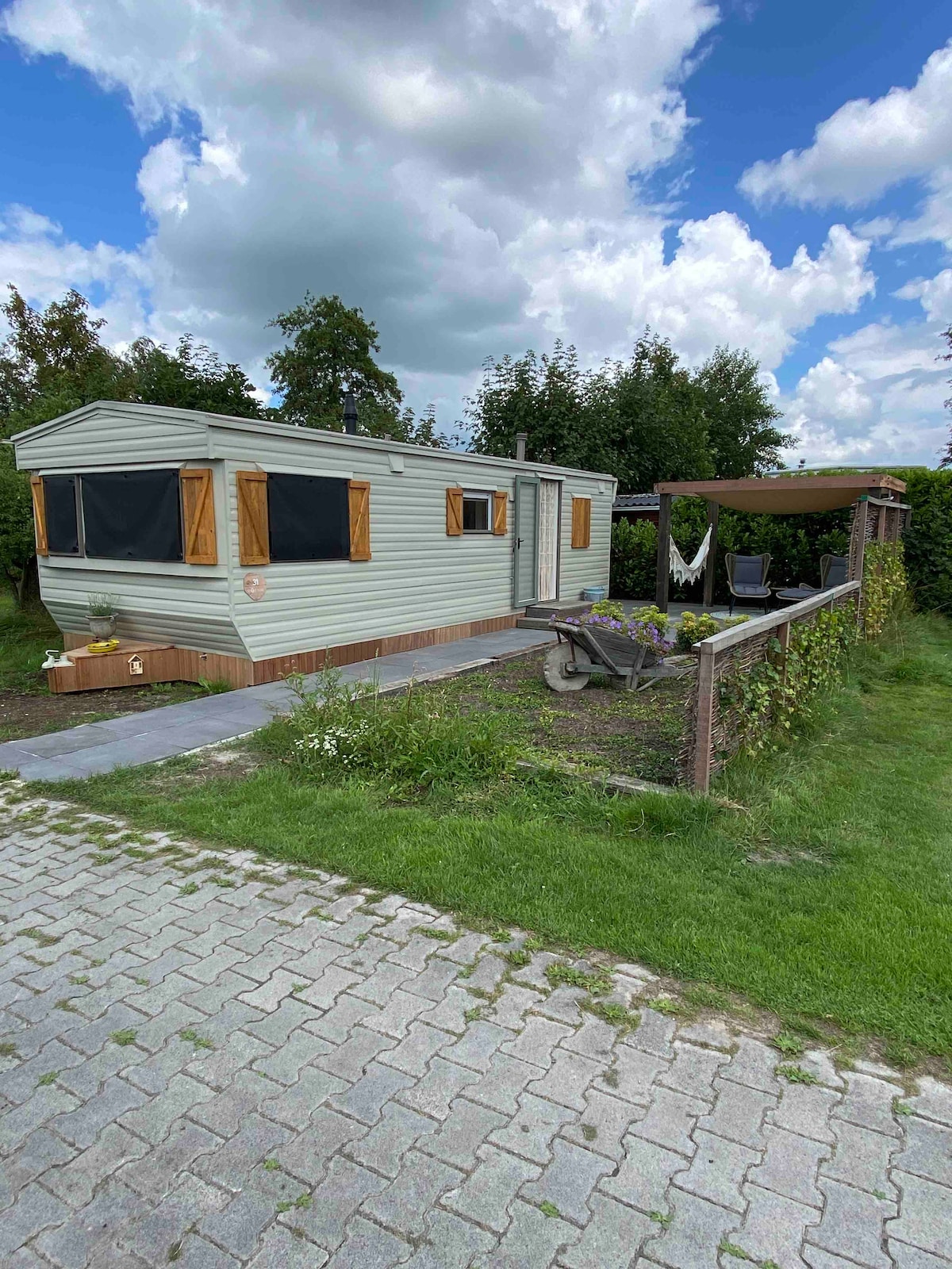 A light green mobile home is featured with wooden window shutters, set within a grassy area. A stone pathway leads to the entrance, and a shaded seating area with chairs is visible beside the home. Surrounding greenery contributes to a tranquil outdoor environment.