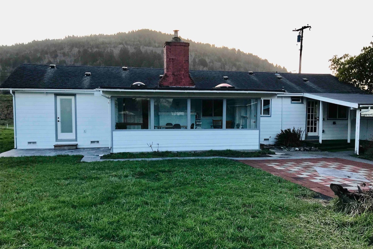 A mid-century lodge-style ranch house is shown with a white exterior and a spacious front yard. Large windows create a bright sunroom, while a brick pathway leads towards the entrance. The backdrop features rolling hills and a soft evening light.
