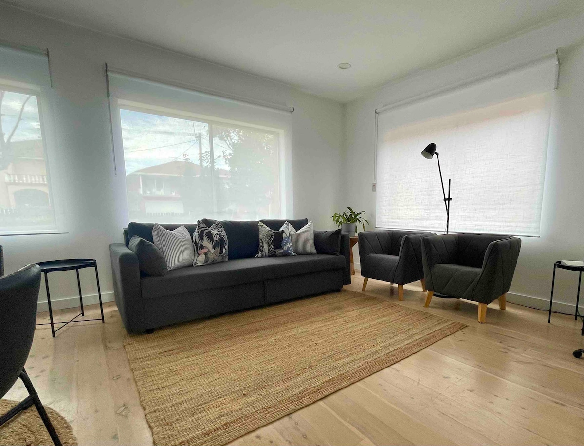 A bright living area features a dark sofa adorned with patterned cushions, complemented by two stylish armchairs. Natural light is filtered through large windows, casting a soft glow on the light wood flooring, while a textured area rug adds warmth to the space.