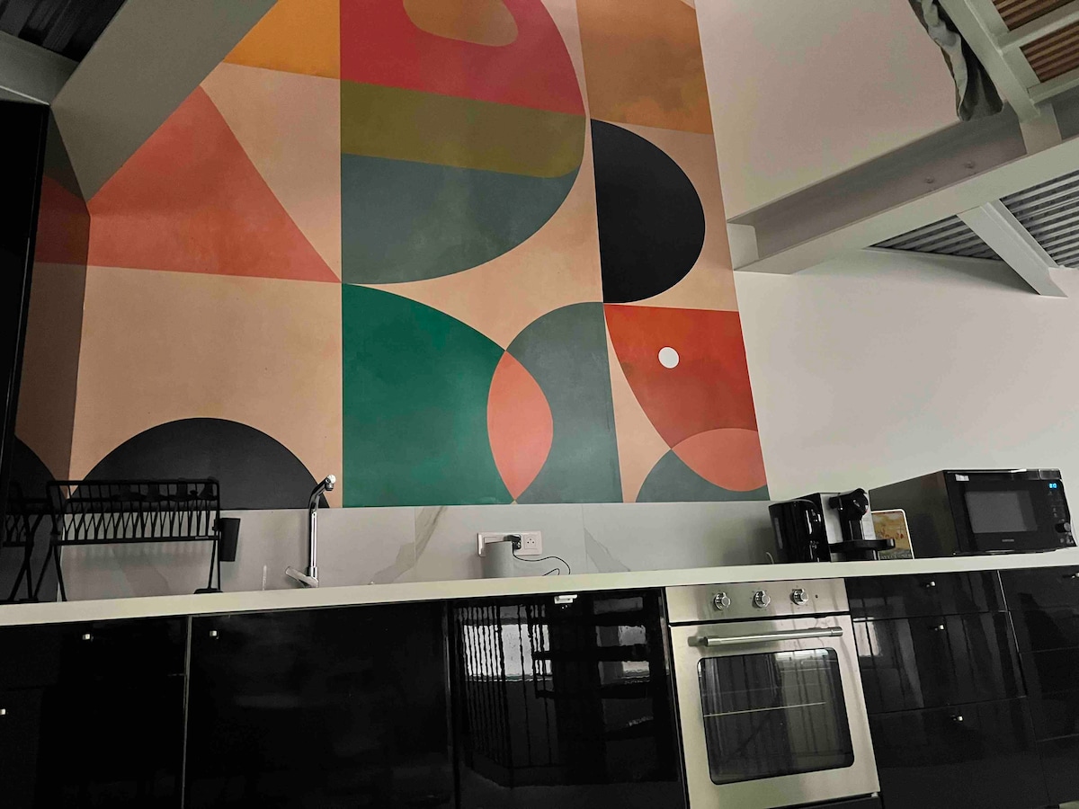 A modern kitchen is highlighted by a large abstract mural featuring shapes in shades of green, red, and black. The sleek, dark cabinets contrast with stainless steel appliances, including an oven and a microwave. A minimalist sink is visible, enhancing the kitchen's contemporary design.