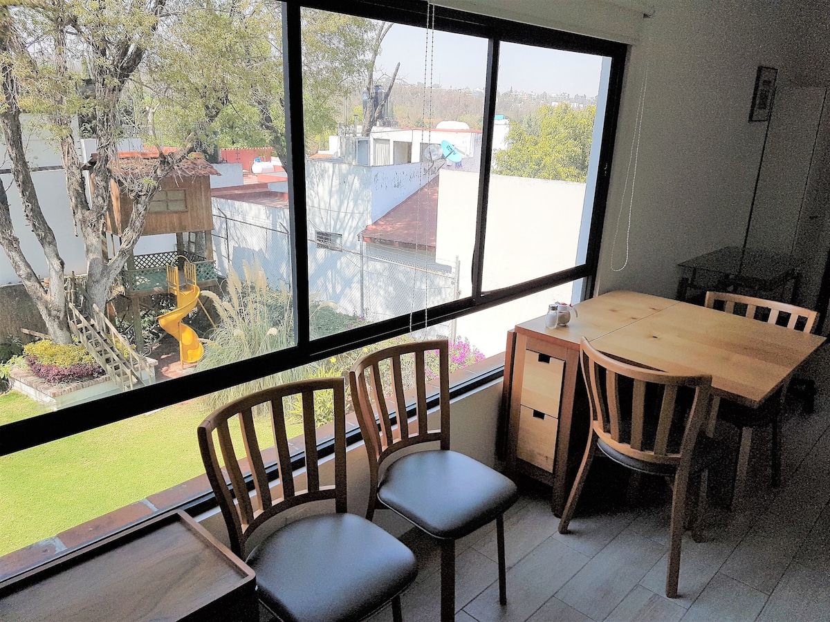 A bright dining area features a wooden table and three chairs, positioned beside large windows that overlook a lush garden. The outdoor space includes a treehouse and a playful slide, creating a serene view filled with greenery.
