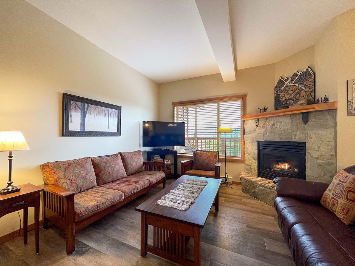 The living area is designed for comfort, featuring a stone fireplace and a mix of seating options, including a leather sofa and patterned chairs. A large window allows natural light to illuminate the space, and a television is positioned for easy viewing.