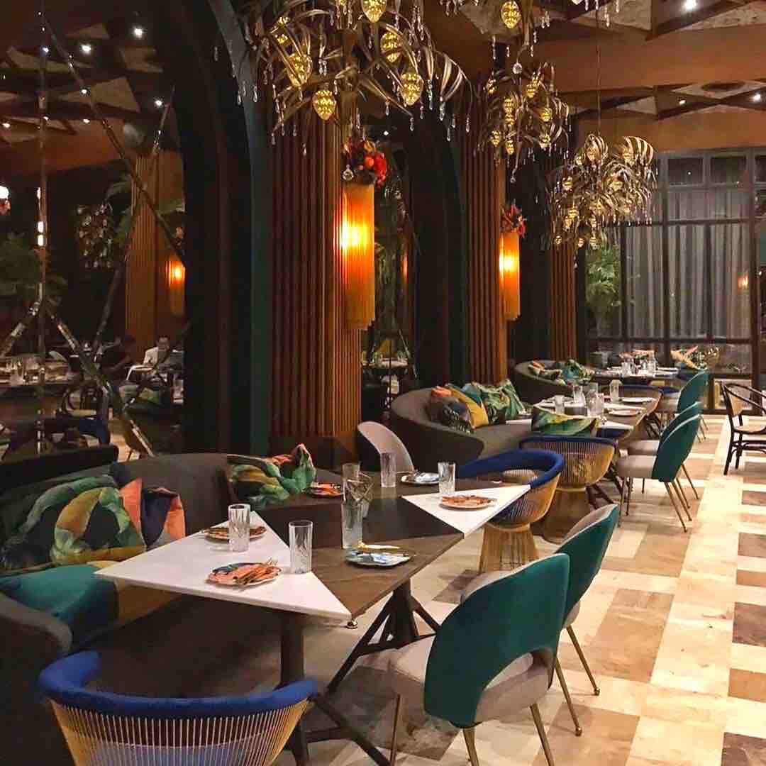 An elegantly designed dining area features plush seating with various colored chairs placed around tables. Ornate light fixtures hang from the ceiling, illuminating the space. Large windows allow natural light to enter, while greenery is visible in the background, enhancing the inviting atmosphere.