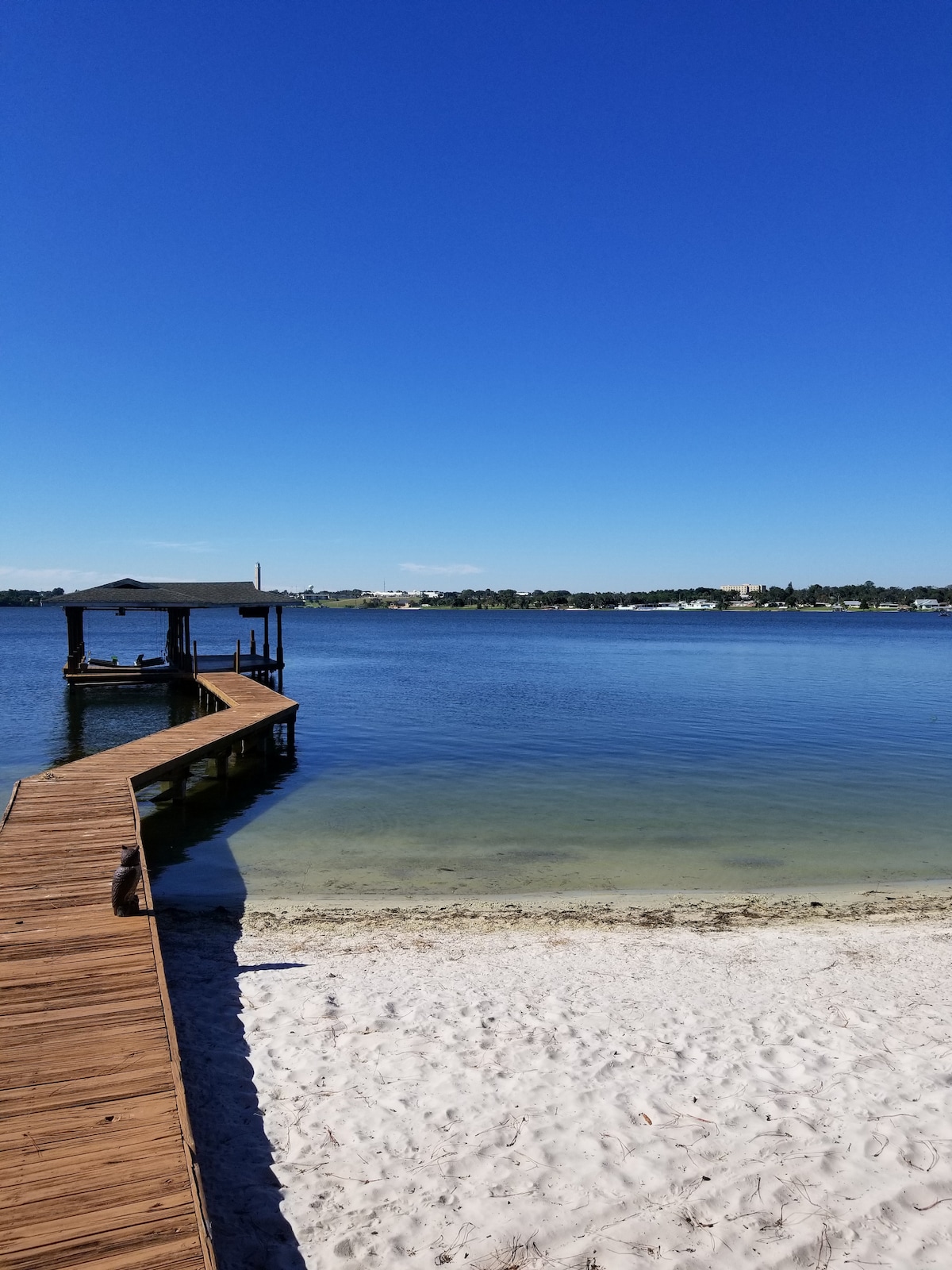 A wooden dock extends over the calm waters of Lake Clay, leading to a covered gazebo. The sandy beachfront is visible in the foreground, while the clear blue sky reflects on the water's surface. Distant scenery includes trees and structures along the shoreline.