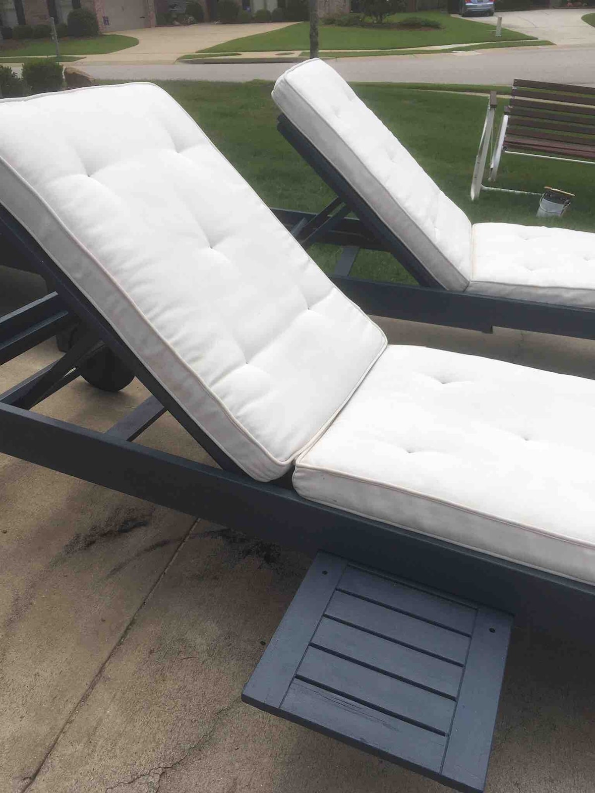 Two adjustable outdoor lounge chairs are positioned to face a grassy area. Each chair features a white cushioned seat with tufted detailing, enhancing comfort. A small wooden platform is attached to each chair for convenience.