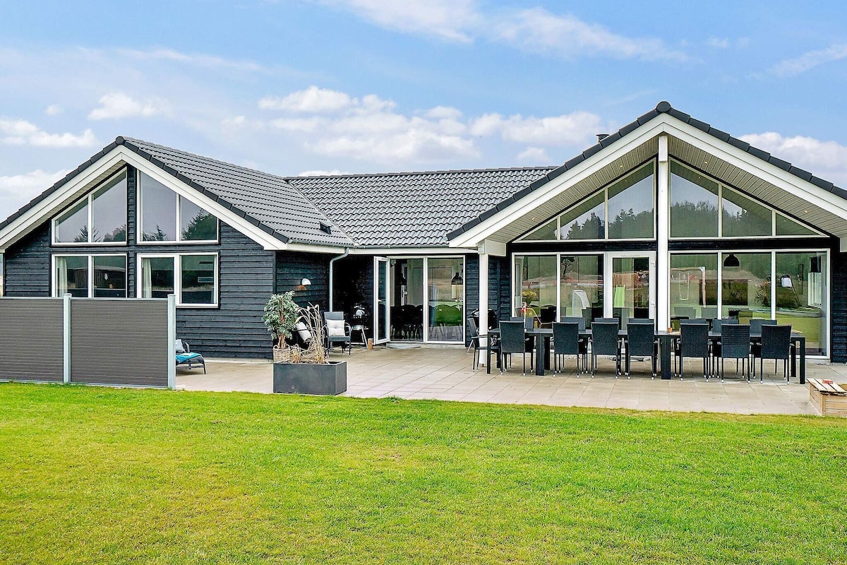 The spacious exterior of the holiday home is showcased, featuring large glass windows allowing for abundant natural light. A covered terrace with a dining table for outdoor meals is visible, along with a well-maintained lawn and distinct architectural lines that highlight the home's modern design.