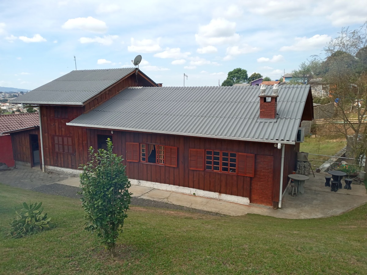 A rustic wooden house, characterized by a sloped metal roof, is set on a grassy slope. Windows with red frames feature on the facade, and a patio area with outdoor seating is visible. Surrounding greenery adds to the peaceful environment.