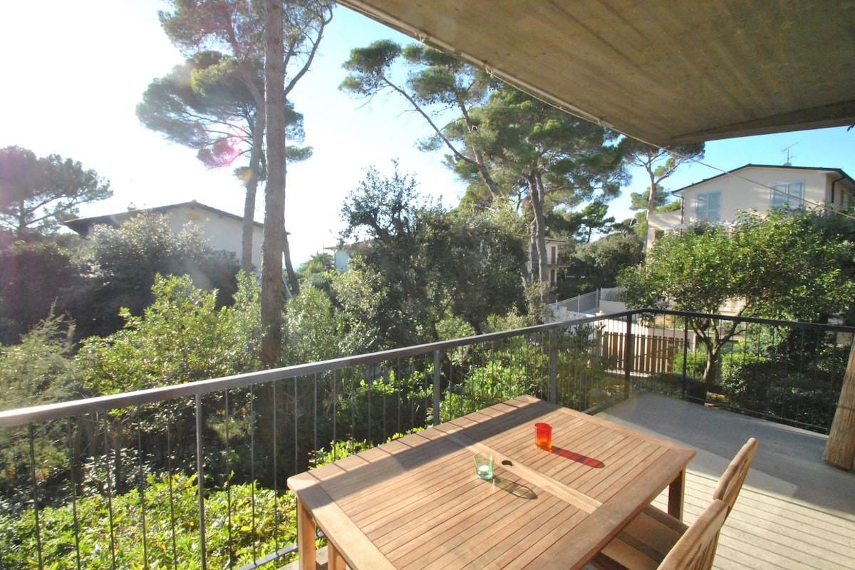A spacious terrace features a wooden dining table, complemented by colorful glassware. Lush greenery surrounds the area, with tall trees providing shade. The image captures a serene outdoor setting, with sunlight gently illuminating the space and distant views of neighboring residences.