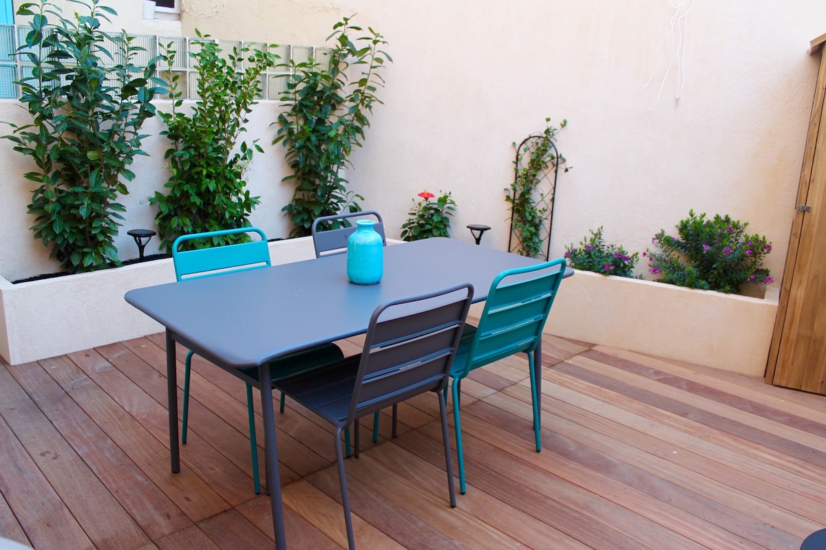 An outdoor terrace is featured, furnished with a rectangular table surrounded by four chairs. Green plants are arranged in the background, enhancing the natural setting. The wooden deck provides a warm, inviting space for relaxation.