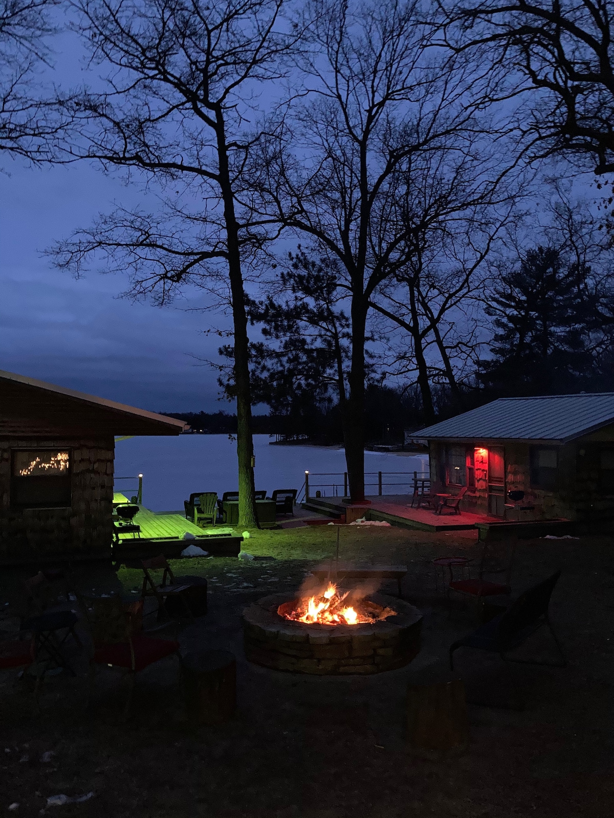A fire pit is surrounded by seating options, with a warm glow from the fire contrasting against the cool twilight. Two cabins are visible, one with red lighting and the other nestled under trees, with a view of the lake in the background.