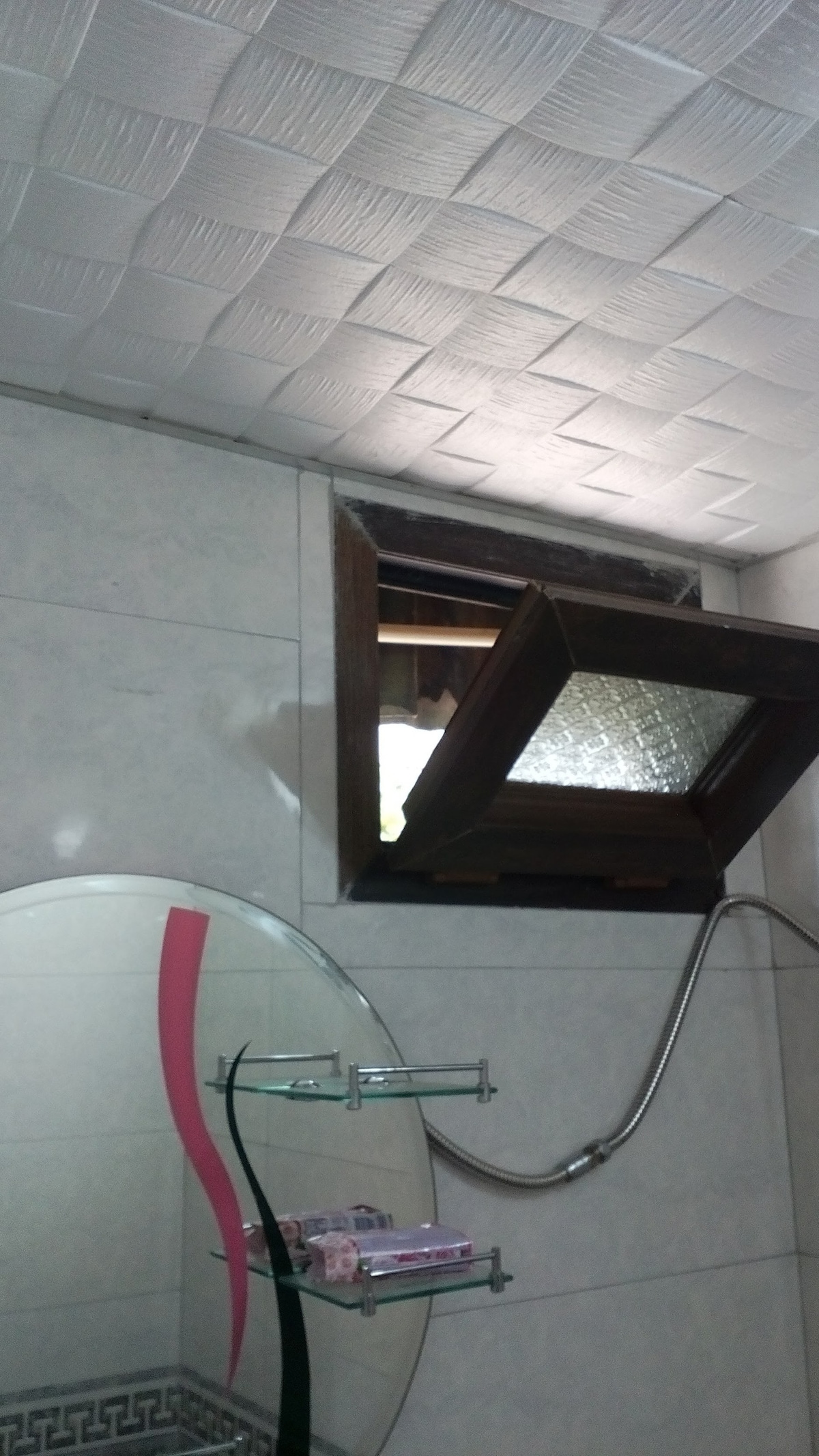 The bathroom features a window with a wooden frame that opens outward, allowing natural light to enter. A round mirror is mounted on the wall, reflecting the white tiled walls and a glass shelf with toiletries. A showerhead is visible in the corner.