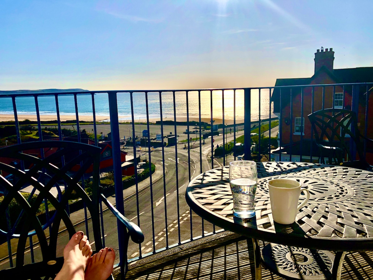 A private balcony is shown, with a table holding a glass of water and a mug. Bare feet rest on the railing, overlooking a sandy beach and distant waves under a bright sky. The sun reflects off the water, illuminating the scene.