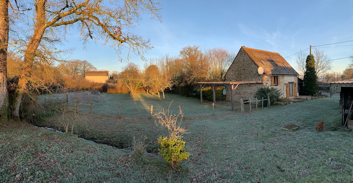A scenic view of the property showcases a converted barn surrounded by frosted grass and trees. Morning light casts warmth on the rustic stone exterior and nearby gardens, which feature a variety of shrubs and an open gazebo area. The landscape offers a tranquil rural setting.
