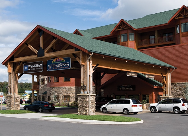 The resort's entrance showcases a grand wooden canopy supported by stone columns, marked by distinctive signage. The exterior features a mix of warm earth tones, with a multi-level structure and a green roof. Several vehicles are parked in front, highlighting accessibility.