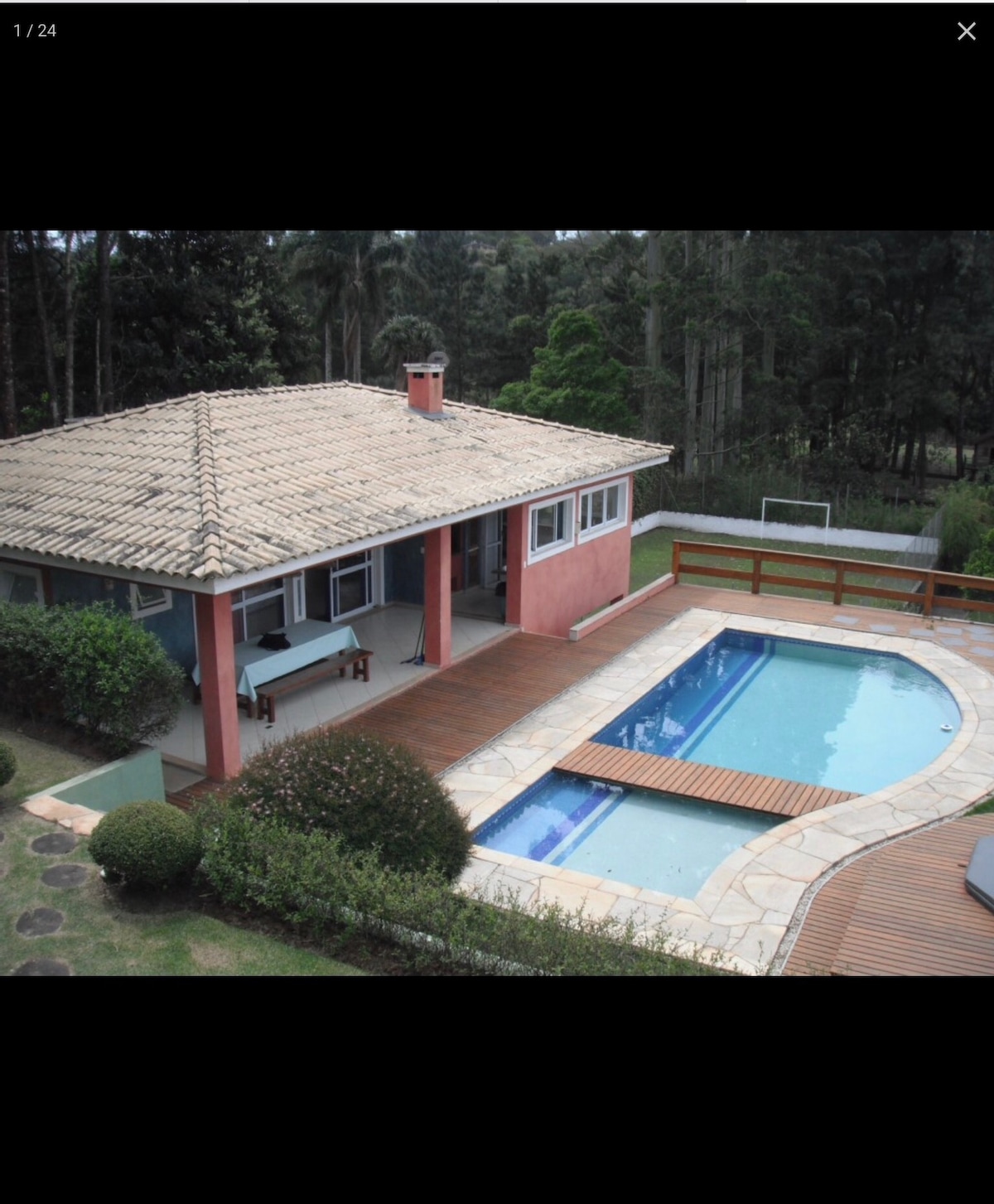 A house with a tiled roof is seen from above, featuring a spacious wooden deck surrounding a rectangular pool. Lush greenery and well-maintained hedges frame the property, while large patio doors provide access to the interior. A dining table is positioned on the deck.