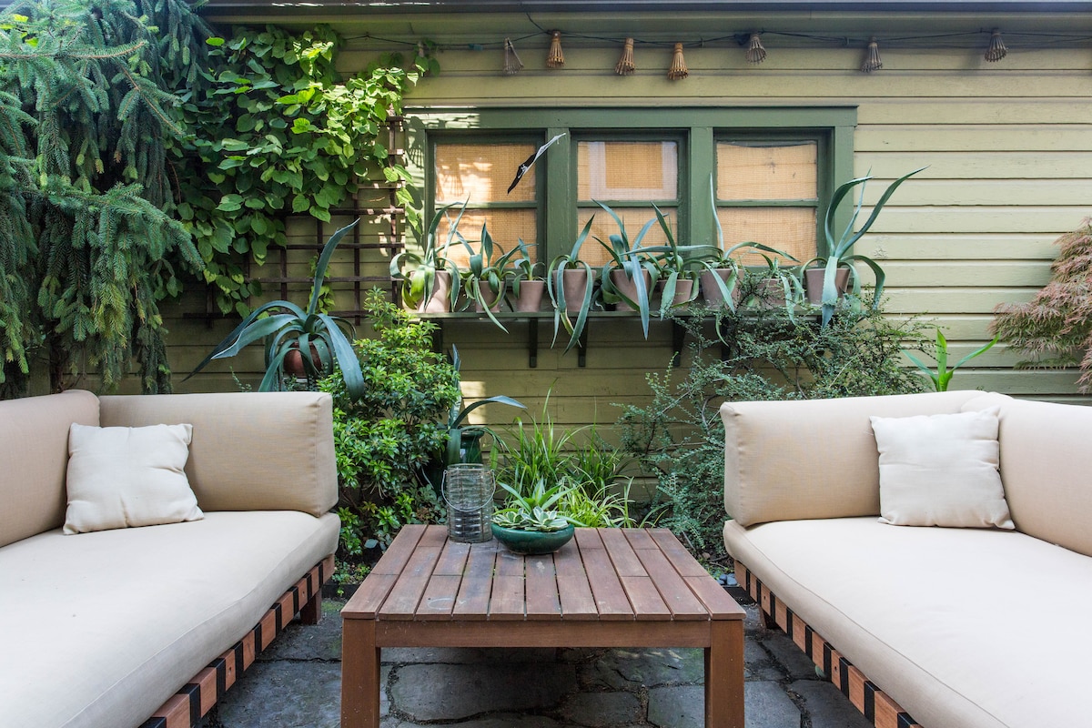 A cozy outdoor seating area features two beige sofas with light-colored cushions, arranged around a wooden coffee table. Lush greenery surrounds the space, including potted plants and climbing vines against a wooden wall, creating a tranquil atmosphere.