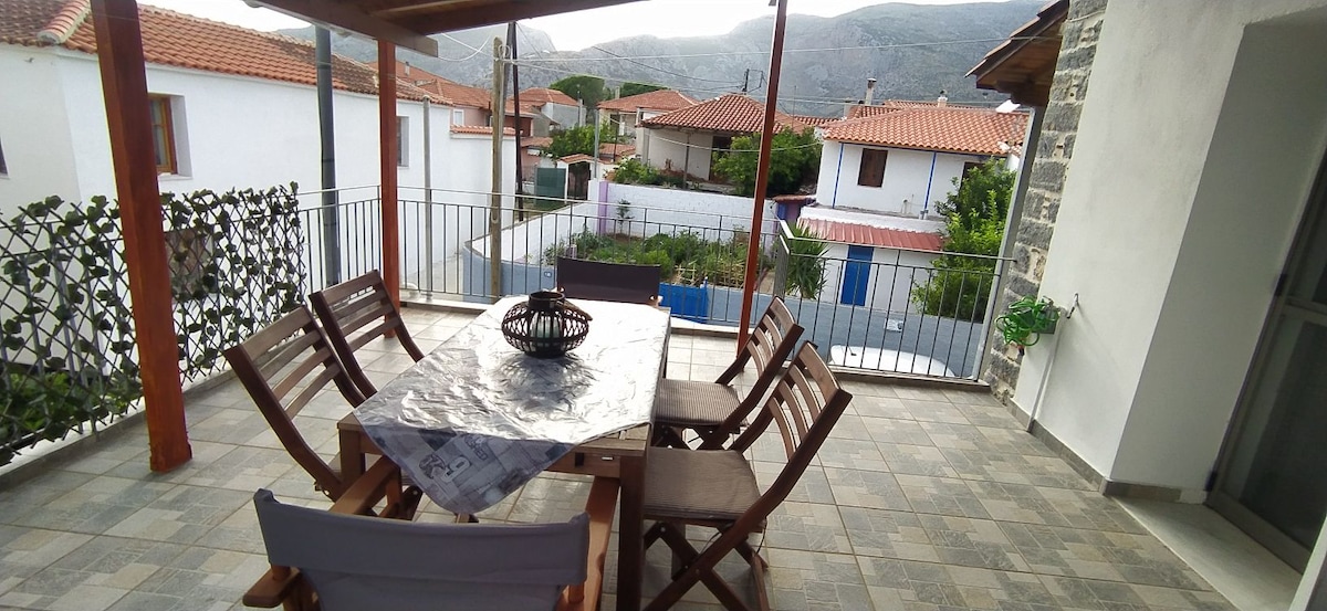 An outdoor dining area is featured, equipped with a large table and several wooden chairs. A canopy provides shade, while a view of neighboring houses and mountains is visible in the background, offering a sense of openness and connection to the surroundings.