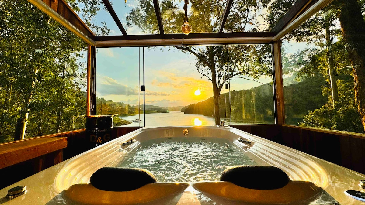 A hot tub with jets is positioned in a glass-enclosed area, offering unobstructed views of the lake and surrounding trees. The sunset casts warm hues across the sky, reflecting on the water, creating a serene atmosphere.