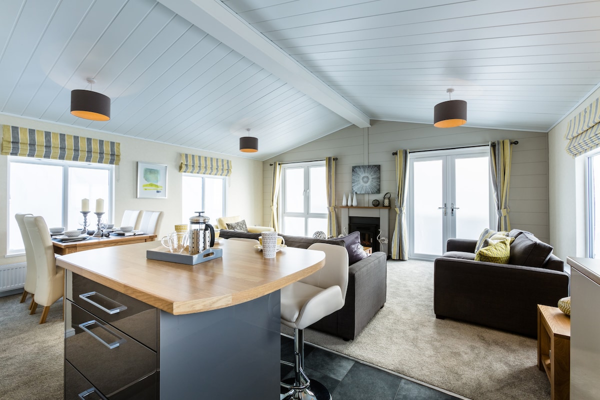 An open-plan living space features a modern kitchen island with seating and a dining area beside large windows. Light fixtures hang from a sloped ceiling, while cozy seating options are arranged around a fireplace, offering a relaxed atmosphere. Natural light fills the room through sliding glass doors.